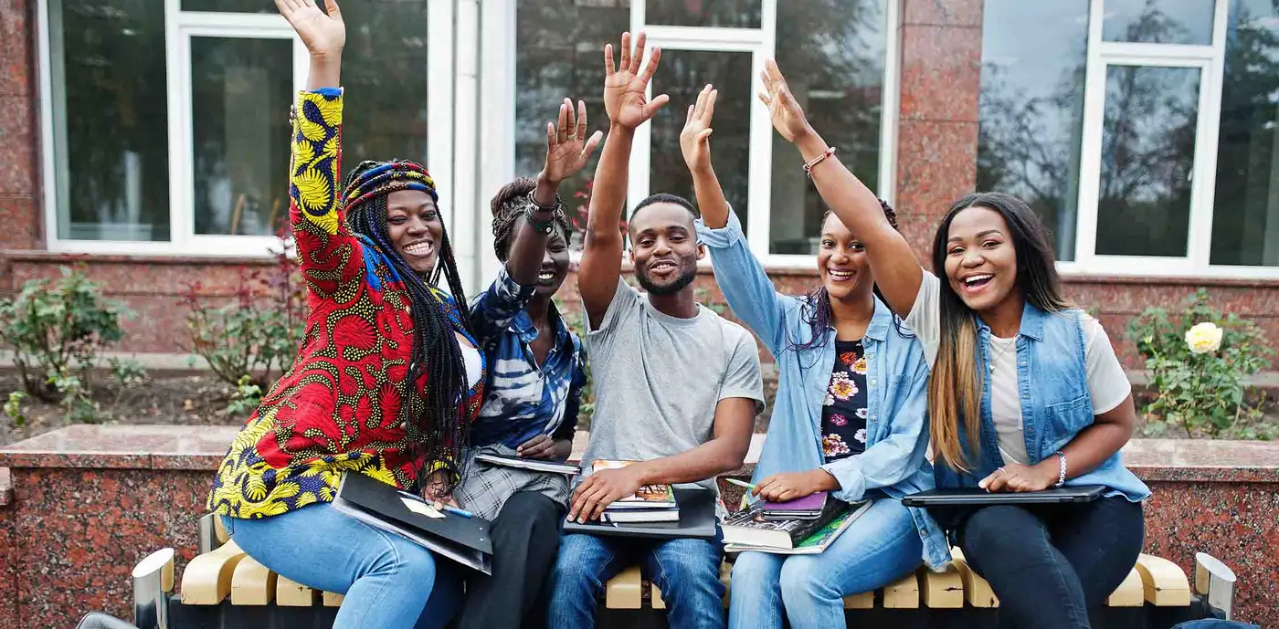 Cinq jeunes adultes assis côte à côte devant un établissement de formation, cahiers et classeurs sur les genoux, le bras levé.